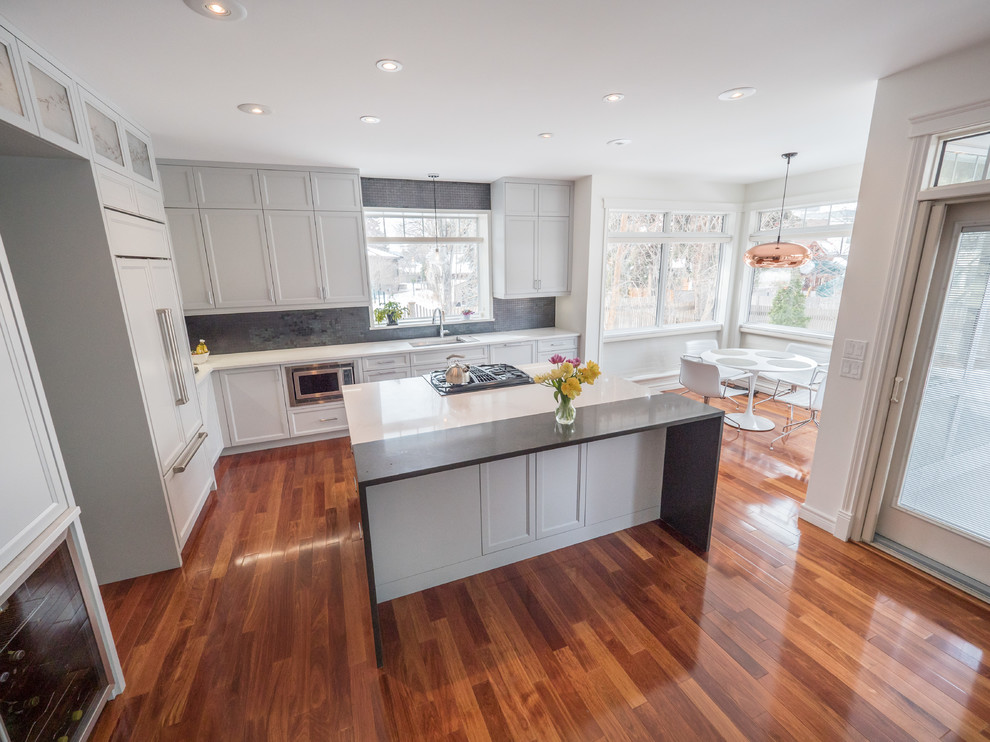 This is an example of a mid-sized contemporary l-shaped open plan kitchen in Edmonton with an undermount sink, white cabinets, quartz benchtops, black splashback, panelled appliances, medium hardwood floors, with island, brown floor, white benchtop and shaker cabinets.