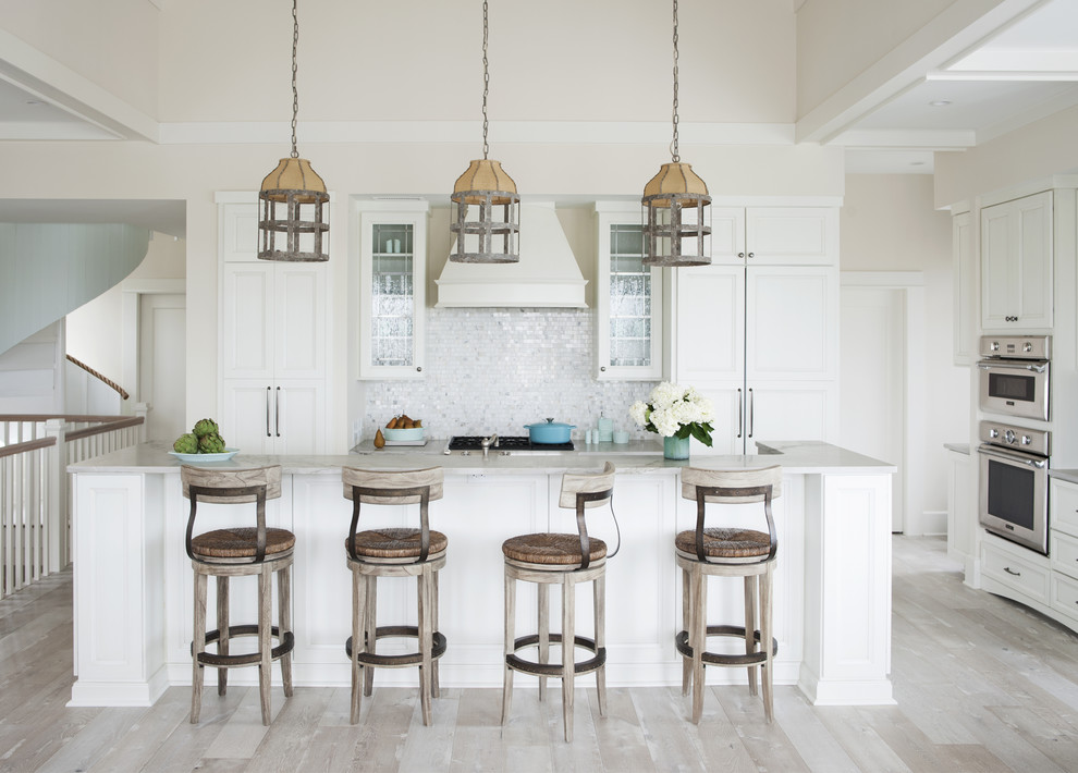 Large beach style l-shaped open plan kitchen in Atlanta with recessed-panel cabinets, white cabinets, stainless steel appliances, light hardwood floors, with island, a farmhouse sink, marble benchtops, white splashback, glass tile splashback and beige floor.