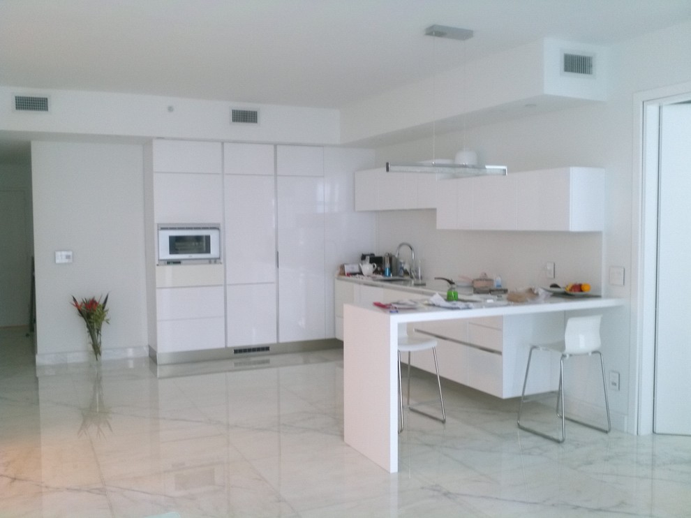 Photo of a small modern l-shaped open plan kitchen in Miami with an undermount sink, flat-panel cabinets, white cabinets, quartzite benchtops, white splashback, stone slab splashback, white appliances and marble floors.