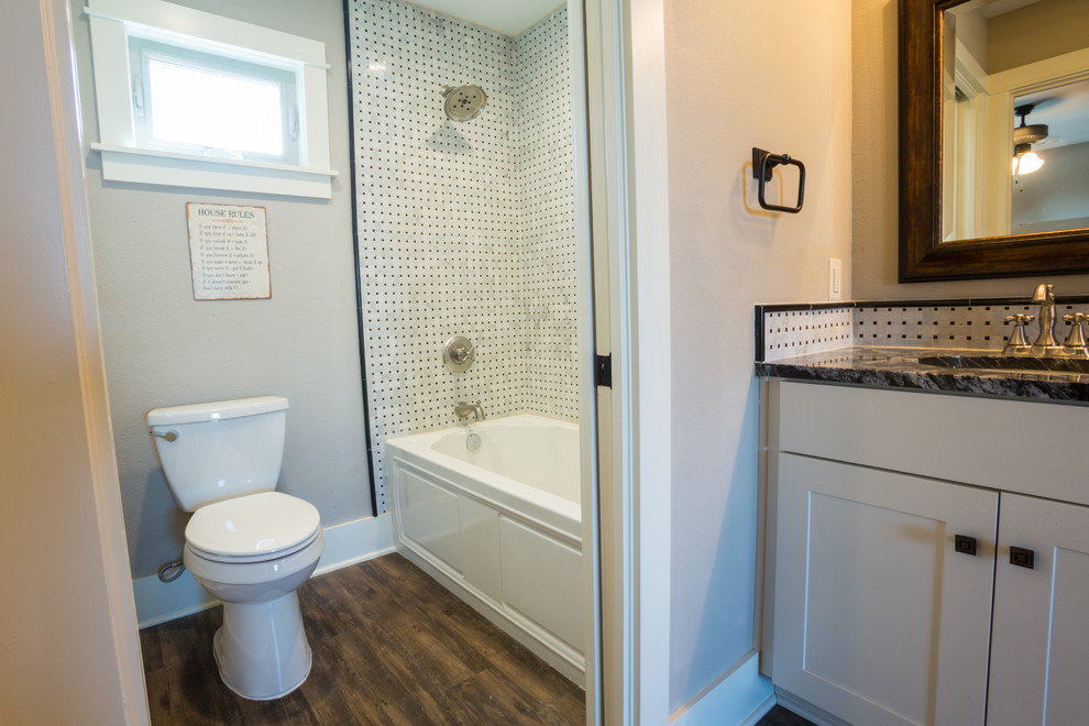 Photo of a mid-sized arts and crafts bathroom in Austin with flat-panel cabinets, white cabinets, granite benchtops, a drop-in tub, a shower/bathtub combo, black tile, ceramic tile, grey walls and dark hardwood floors.