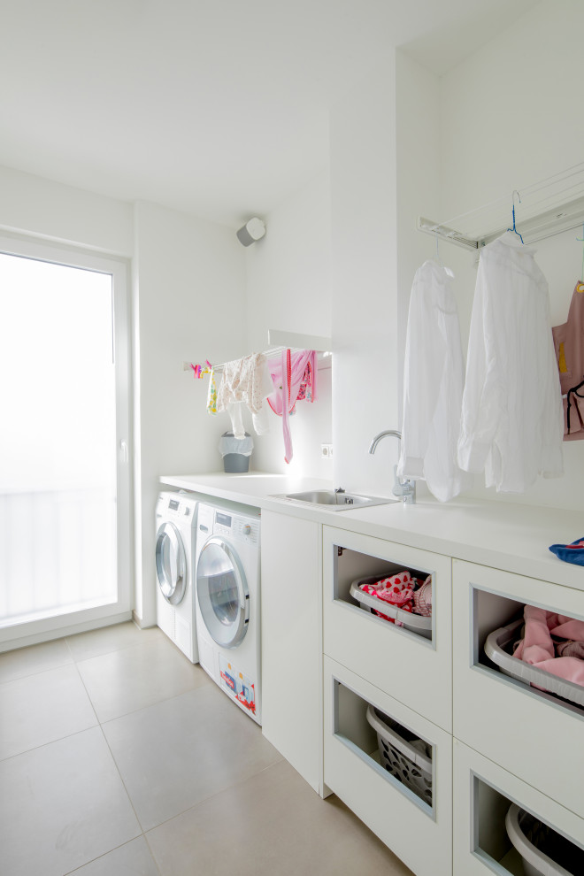 Photo of a mid-sized contemporary single-wall dedicated laundry room in Dusseldorf with a drop-in sink, flat-panel cabinets, white cabinets, wood benchtops, white splashback, white walls, ceramic floors, an integrated washer and dryer, grey floor and white benchtop.