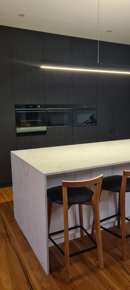 Large contemporary galley open plan kitchen in Geelong with a double-bowl sink, dark wood cabinets, solid surface benchtops, white splashback, ceramic splashback, black appliances, medium hardwood floors, with island and white benchtop.