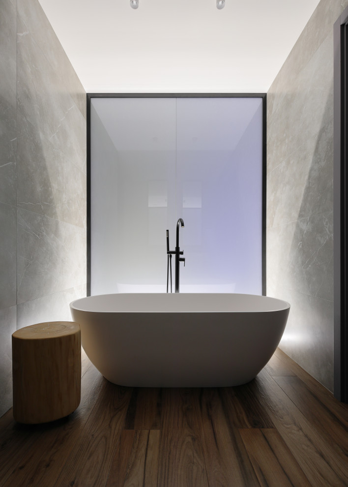 Skandinavisk inredning av ett mellanstort brun brunt en-suite badrum, med släta luckor, grå skåp, ett fristående badkar, en vägghängd toalettstol, grå kakel, porslinskakel, grå väggar, träbänkskiva och brunt golv