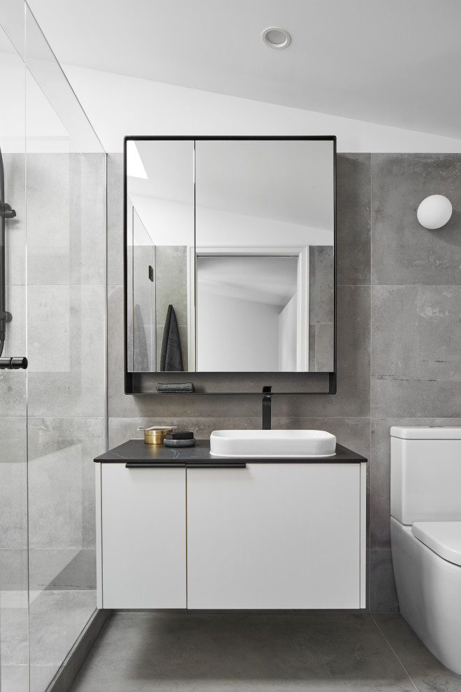 This is an example of a mid-sized contemporary 3/4 bathroom in Melbourne with flat-panel cabinets, a one-piece toilet, gray tile, ceramic tile, grey walls, ceramic floors, a vessel sink, solid surface benchtops, grey floor, a hinged shower door, black benchtops, a single vanity, a floating vanity, vaulted and white cabinets.
