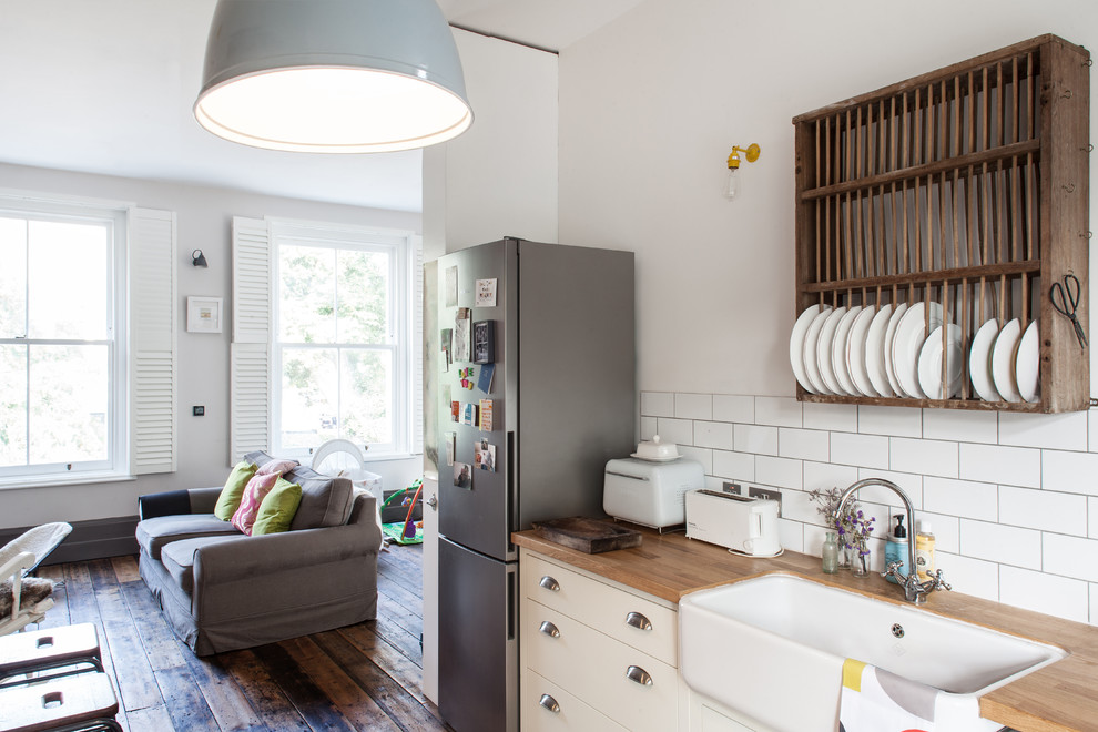 Small traditional l-shaped open plan kitchen in London with a farmhouse sink, flat-panel cabinets, white cabinets, wood benchtops, white splashback, subway tile splashback, stainless steel appliances, a peninsula and medium hardwood floors.