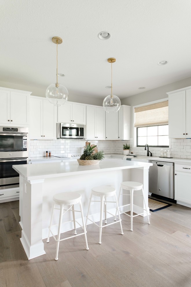 Design ideas for a beach style l-shaped kitchen in Miami with a farmhouse sink, shaker cabinets, white cabinets, white splashback, subway tile splashback, light hardwood floors, with island, beige floor and white benchtop.