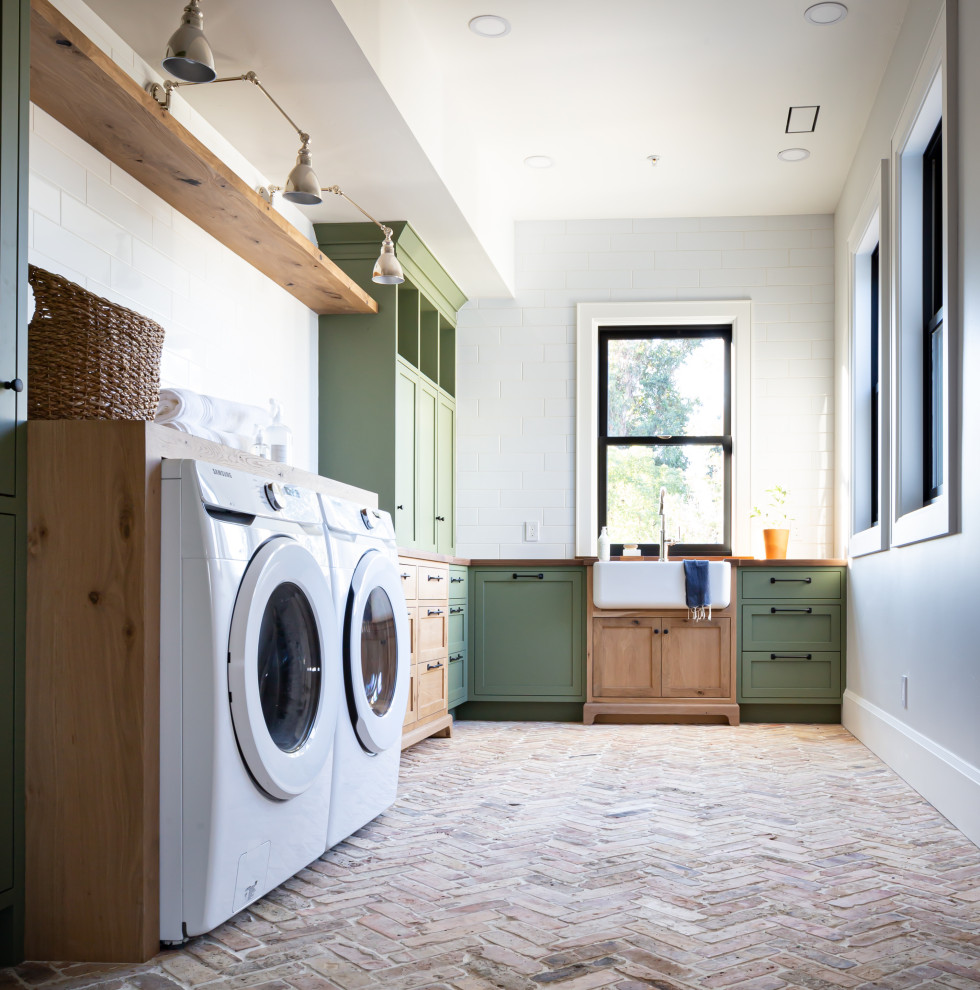 Large rural l-shaped separated utility room in San Francisco with a belfast sink, shaker cabinets, green cabinets, wood worktops, white splashback, porcelain splashback, white walls, brick flooring, an integrated washer and dryer, beige floors and brown worktops.
