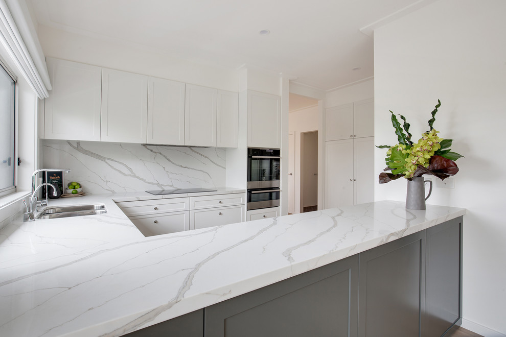 This is an example of a large contemporary u-shaped open plan kitchen in Sydney with a drop-in sink, shaker cabinets, white cabinets, quartz benchtops, white splashback, stone slab splashback, white appliances, medium hardwood floors, no island, beige floor and multi-coloured benchtop.