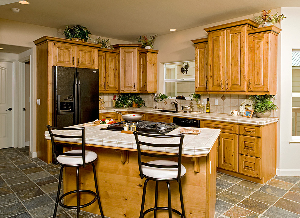 Eat-in kitchen - small rustic l-shaped slate floor eat-in kitchen idea with an undermount sink, raised-panel cabinets, medium tone wood cabinets, tile countertops, beige backsplash, porcelain backsplash, black appliances and an island