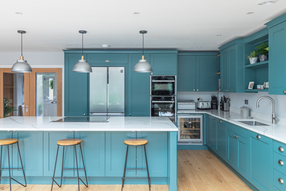Large classic l-shaped open plan kitchen in Hertfordshire with a built-in sink, shaker cabinets, green cabinets, quartz worktops, integrated appliances, laminate floors, an island and white worktops.