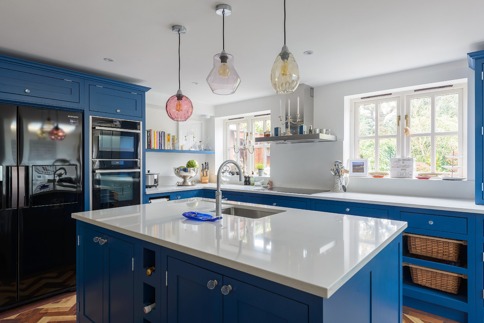 This is an example of a mid-sized traditional l-shaped eat-in kitchen in Surrey with an integrated sink, shaker cabinets, blue cabinets, solid surface benchtops, white splashback, black appliances, medium hardwood floors and with island.