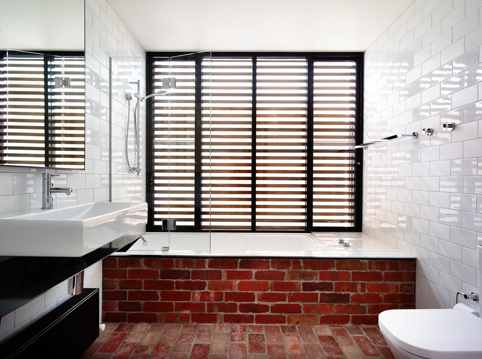 This is an example of a mid-sized contemporary bathroom in Melbourne with white tile, subway tile, black cabinets, an open shower, a wall-mount toilet, white walls, stainless steel benchtops, brick floors, an alcove tub, a trough sink, red floor and an open shower.