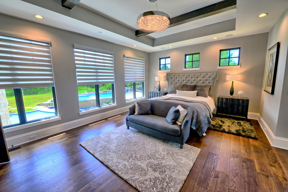 Diseño de dormitorio principal grande con paredes grises, suelo de madera en tonos medios, chimenea lineal, marco de chimenea de piedra, suelo marrón y vigas vistas