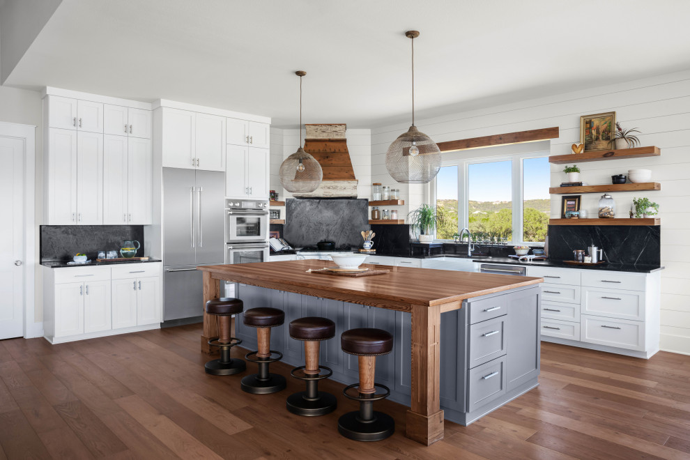 This is an example of a large beach style l-shaped eat-in kitchen in Austin with a farmhouse sink, shaker cabinets, grey cabinets, black splashback, stone slab splashback, stainless steel appliances, medium hardwood floors, with island, brown floor, black benchtop and wood benchtops.