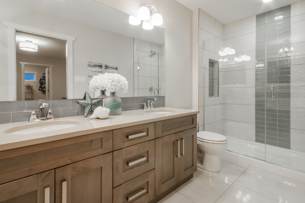 This is an example of a mid-sized contemporary master bathroom in Calgary with shaker cabinets, medium wood cabinets, a drop-in tub, an alcove shower, a one-piece toilet, gray tile, white tile, porcelain tile, white walls, porcelain floors, an undermount sink and solid surface benchtops.