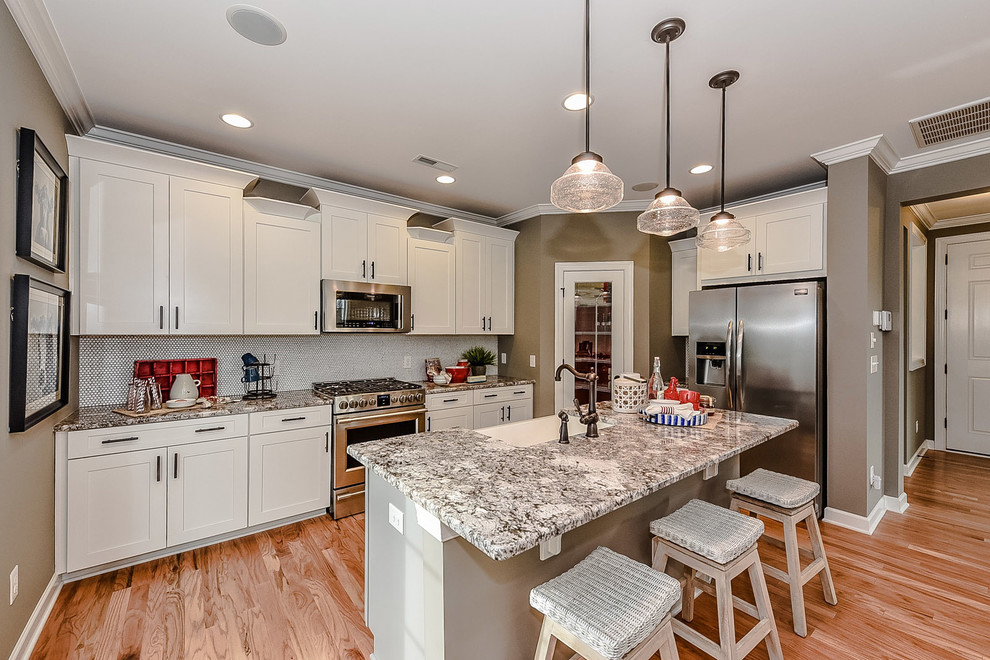 Photo of a small country single-wall open plan kitchen in Charlotte with a farmhouse sink, recessed-panel cabinets, white cabinets, granite benchtops, grey splashback, mosaic tile splashback, stainless steel appliances, light hardwood floors and with island.