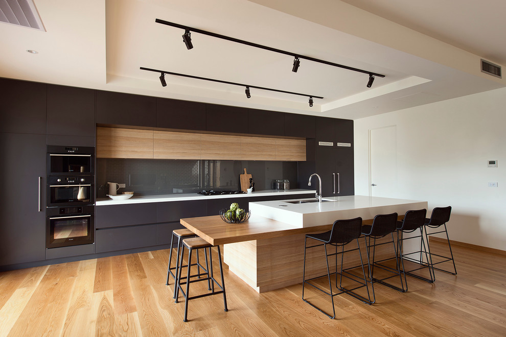 Design ideas for a contemporary kitchen in Los Angeles with an undermount sink, flat-panel cabinets, black cabinets, black splashback, glass sheet splashback, black appliances, light hardwood floors, with island, white benchtop and recessed.