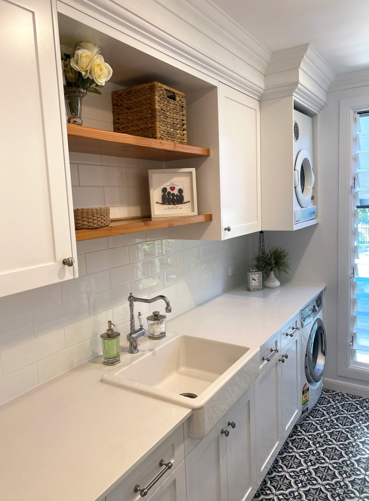 This is an example of a large beach style galley dedicated laundry room in Brisbane with a farmhouse sink, shaker cabinets, white cabinets, quartz benchtops, white splashback, subway tile splashback, white walls, ceramic floors, black floor, white benchtop, recessed and panelled walls.