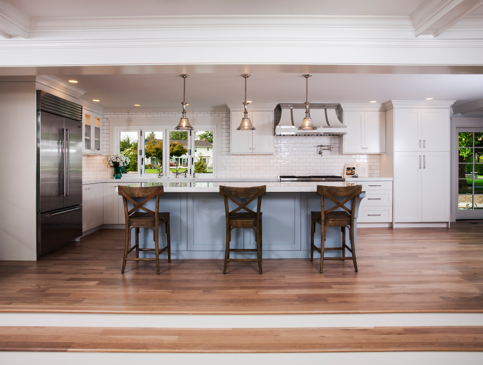 This is an example of a large traditional l-shaped open plan kitchen in Seattle with a farmhouse sink, shaker cabinets, white cabinets, quartz benchtops, white splashback, subway tile splashback, stainless steel appliances, light hardwood floors, with island, beige floor, white benchtop and exposed beam.