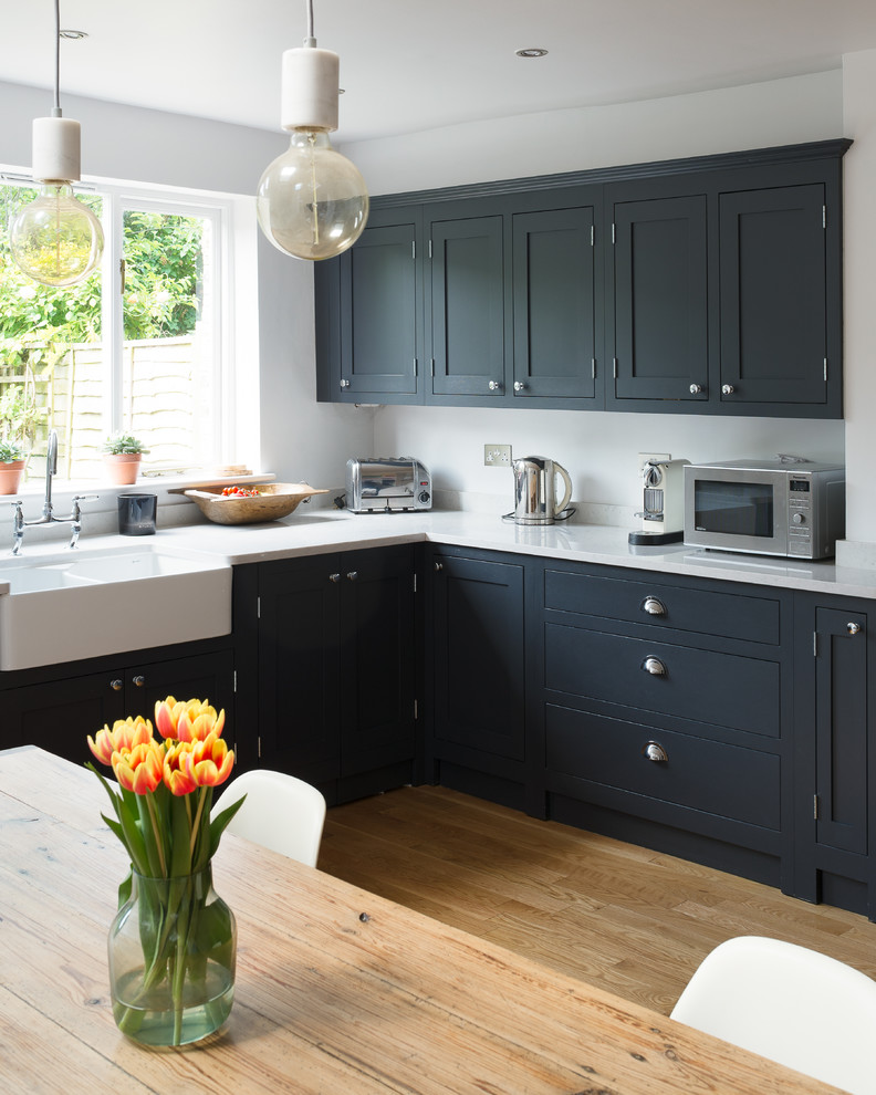 This is an example of a small country eat-in kitchen in London with shaker cabinets, light hardwood floors, a farmhouse sink and black cabinets.