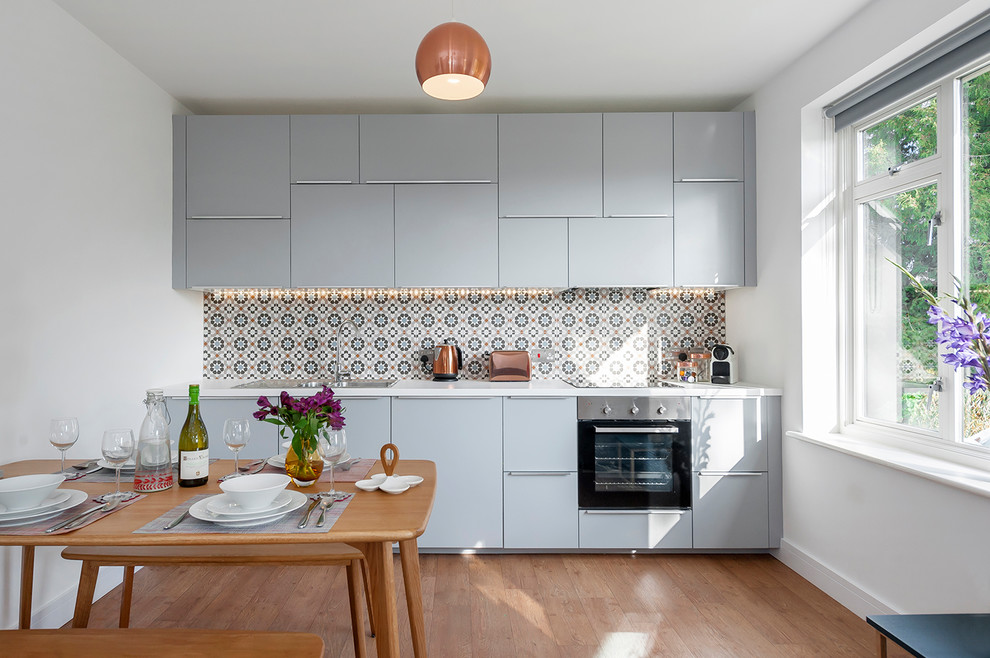 Small contemporary single-wall eat-in kitchen in Wiltshire with flat-panel cabinets, blue cabinets, stainless steel appliances, light hardwood floors, brown floor, a drop-in sink and multi-coloured splashback.