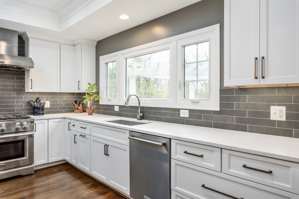 This is an example of a large contemporary eat-in kitchen in DC Metro with an undermount sink, recessed-panel cabinets, white cabinets, quartz benchtops, grey splashback, ceramic splashback, stainless steel appliances, dark hardwood floors, with island, brown floor and white benchtop.