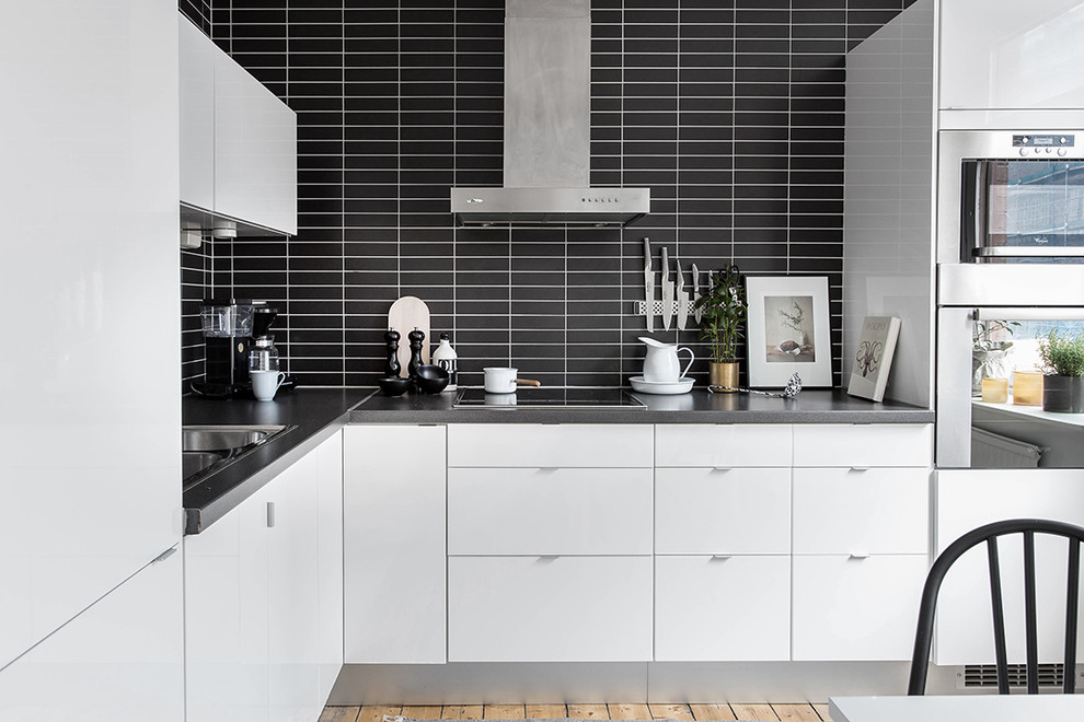 Inspiration for a mid-sized scandinavian l-shaped kitchen in Gothenburg with a drop-in sink, flat-panel cabinets, white cabinets, black splashback, stainless steel appliances, granite benchtops and light hardwood floors.