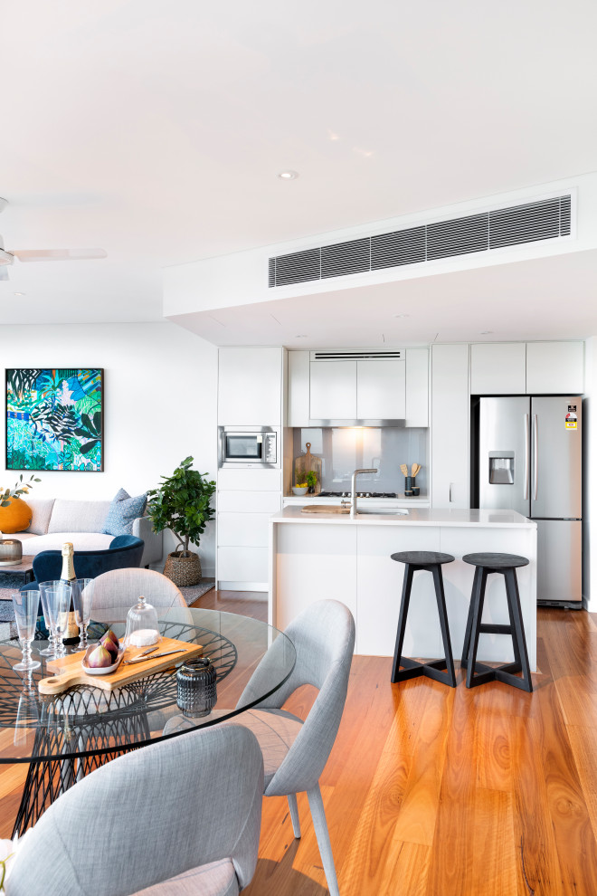 This is an example of a small contemporary galley open plan kitchen in Brisbane with an undermount sink, flat-panel cabinets, white cabinets, grey splashback, glass sheet splashback, stainless steel appliances, with island, brown floor and white benchtop.