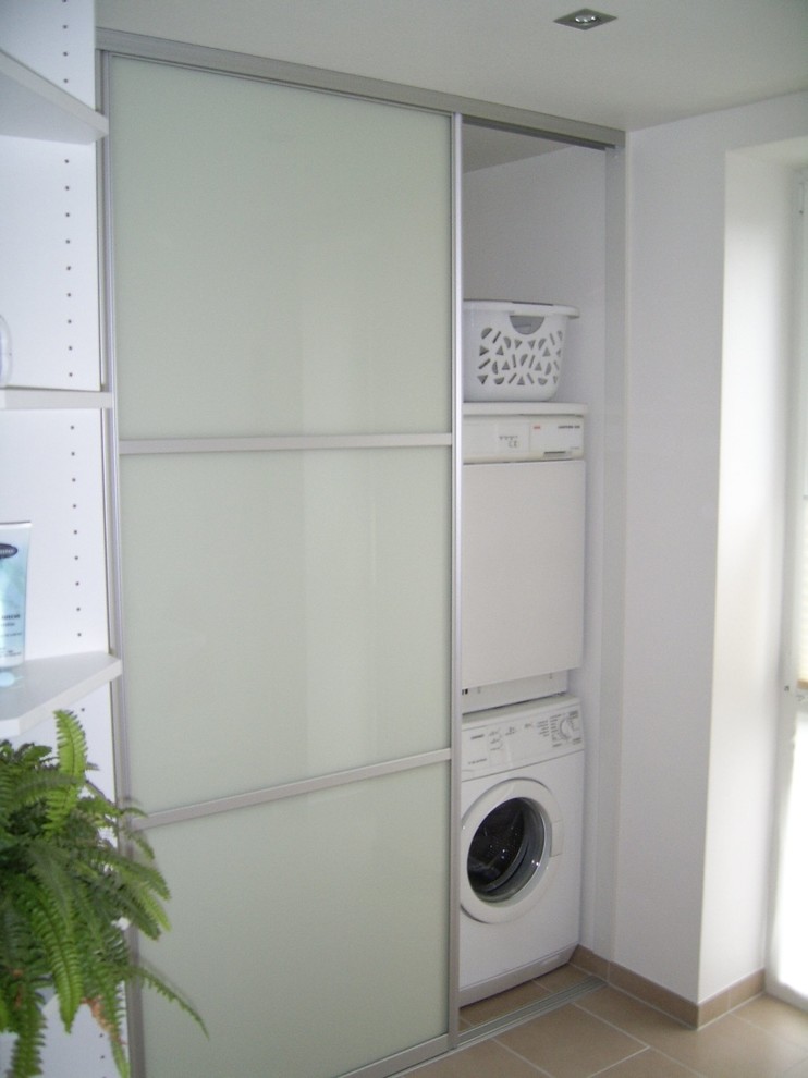 Mid-sized contemporary single-wall laundry cupboard in Cologne with a drop-in sink, glass-front cabinets, white cabinets, wood benchtops, white walls, porcelain floors, a stacked washer and dryer and beige floor.