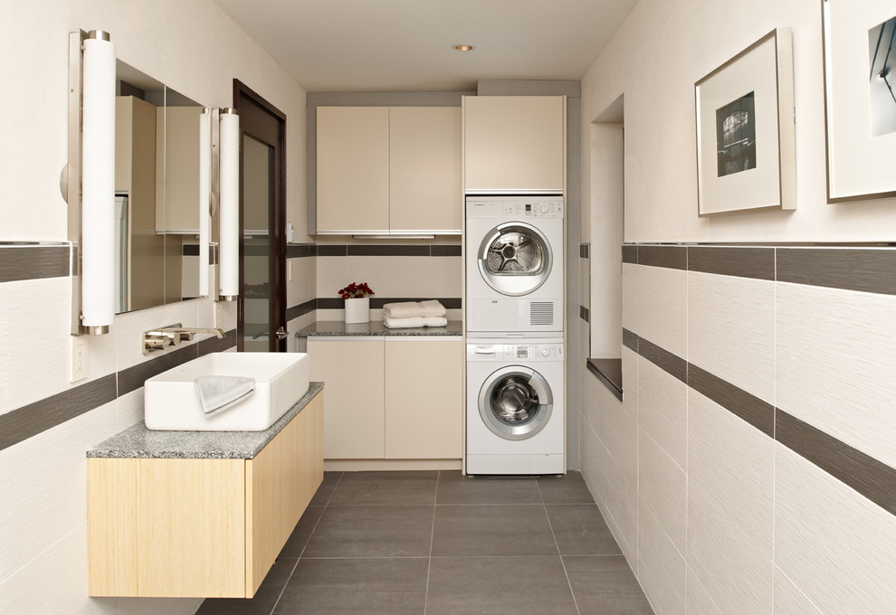 Photo of a contemporary l-shaped utility room in Minneapolis with flat-panel cabinets, beige cabinets, a stacked washer and dryer, grey floor and grey benchtop.