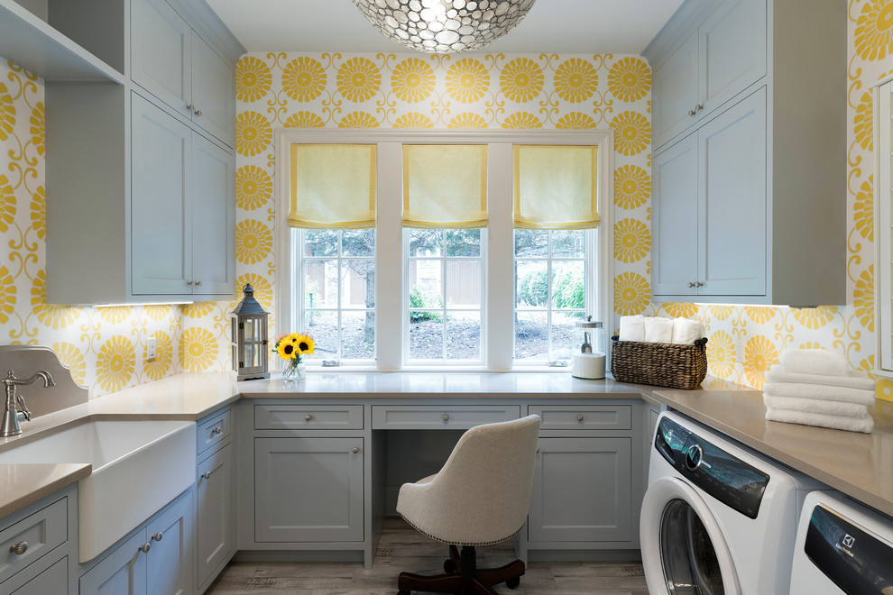 This is an example of a traditional u-shaped utility room in Minneapolis with a farmhouse sink, recessed-panel cabinets, solid surface benchtops, yellow walls, a side-by-side washer and dryer, grey floor and grey cabinets.