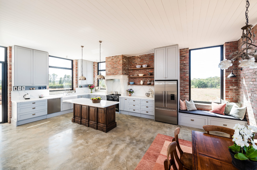 This is an example of an expansive country l-shaped open plan kitchen in Melbourne with a farmhouse sink, shaker cabinets, grey cabinets, quartz benchtops, white splashback, marble splashback, stainless steel appliances, concrete floors, with island, grey floor and white benchtop.