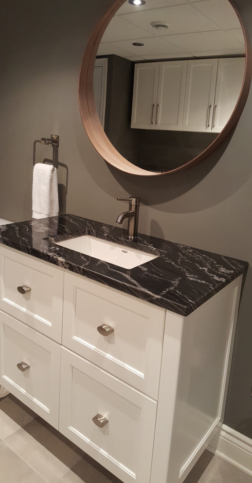 Photo of a mid-sized contemporary galley utility room in Montreal with an undermount sink, shaker cabinets, white cabinets, granite benchtops, grey walls, porcelain floors, a side-by-side washer and dryer, grey floor and black benchtop.