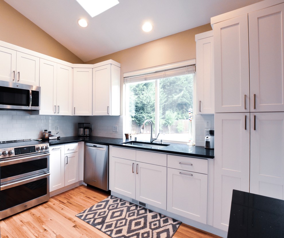 Mid-sized traditional u-shaped eat-in kitchen in Seattle with granite benchtops, white splashback, ceramic splashback, stainless steel appliances, light hardwood floors, black benchtop, an integrated sink, shaker cabinets, white cabinets and with island.