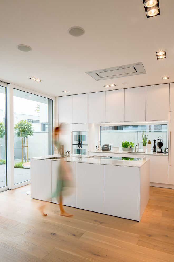 Large modern galley open plan kitchen in Dusseldorf with an undermount sink, flat-panel cabinets, white cabinets, solid surface benchtops, white splashback, glass sheet splashback, white appliances, medium hardwood floors, with island, brown floor and white benchtop.