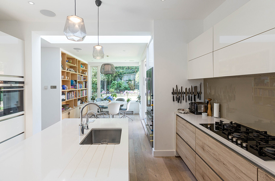 Inspiration for a mid-sized contemporary u-shaped open plan kitchen in London with an integrated sink, flat-panel cabinets, light wood cabinets, solid surface benchtops, brown splashback, glass sheet splashback, black appliances, light hardwood floors, with island, beige floor and grey benchtop.