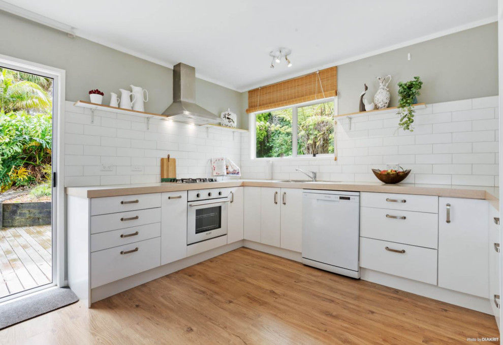 Photo of a mid-sized midcentury u-shaped eat-in kitchen in Auckland with white cabinets, white splashback, ceramic splashback, white appliances, light hardwood floors, no island and brown floor.
