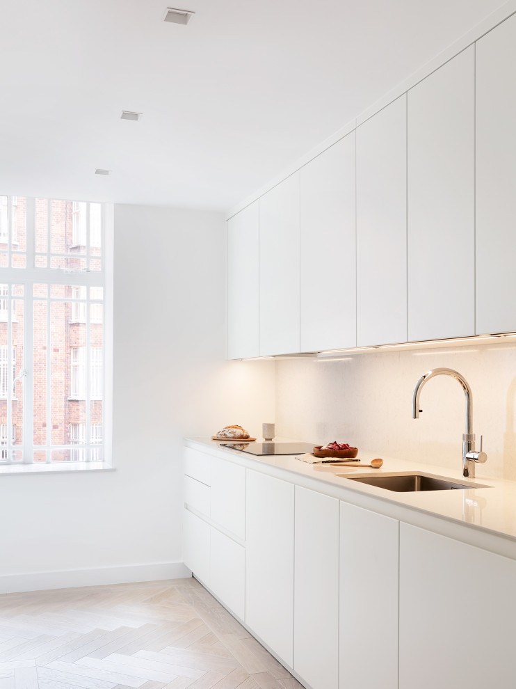 Inspiration for a small contemporary galley separate kitchen in London with a single-bowl sink, flat-panel cabinets, beige cabinets, solid surface benchtops, beige splashback, black appliances, light hardwood floors, beige floor and beige benchtop.