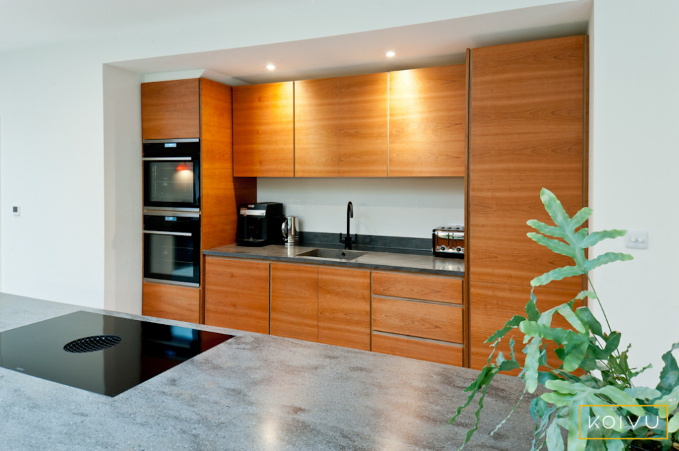 Large contemporary single-wall open plan kitchen in Kent with a drop-in sink, flat-panel cabinets, medium wood cabinets, solid surface benchtops, black appliances, porcelain floors, with island, grey floor and grey benchtop.