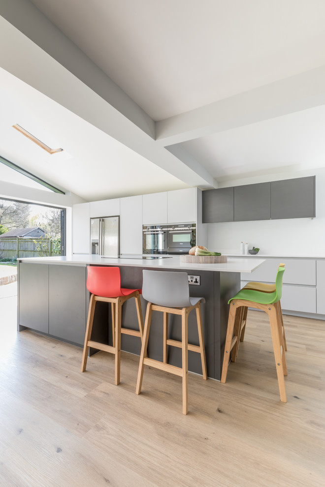 This is an example of a medium sized contemporary galley open plan kitchen in Sussex with an integrated sink, flat-panel cabinets, grey cabinets, composite countertops, stainless steel appliances, an island and white worktops.