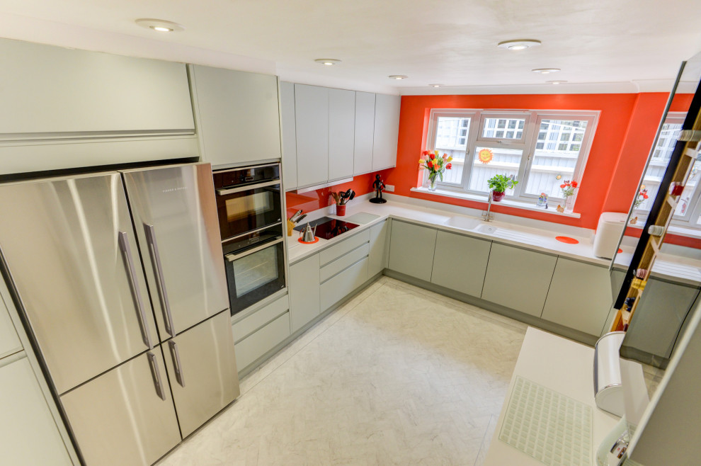 Design ideas for a medium sized nautical grey and white l-shaped enclosed kitchen in Sussex with a built-in sink, flat-panel cabinets, green cabinets, composite countertops, orange splashback, glass sheet splashback, black appliances, lino flooring, a breakfast bar, white floors, white worktops and feature lighting.