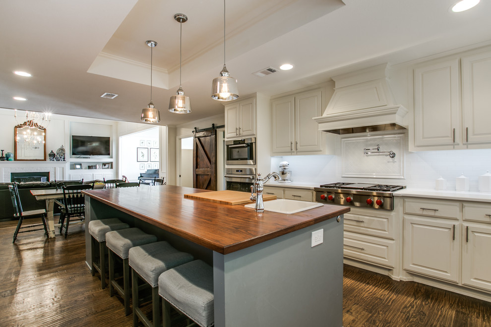 This is an example of a large traditional u-shaped eat-in kitchen in Dallas with a farmhouse sink, raised-panel cabinets, white cabinets, wood benchtops, white splashback, ceramic splashback, stainless steel appliances, medium hardwood floors and with island.