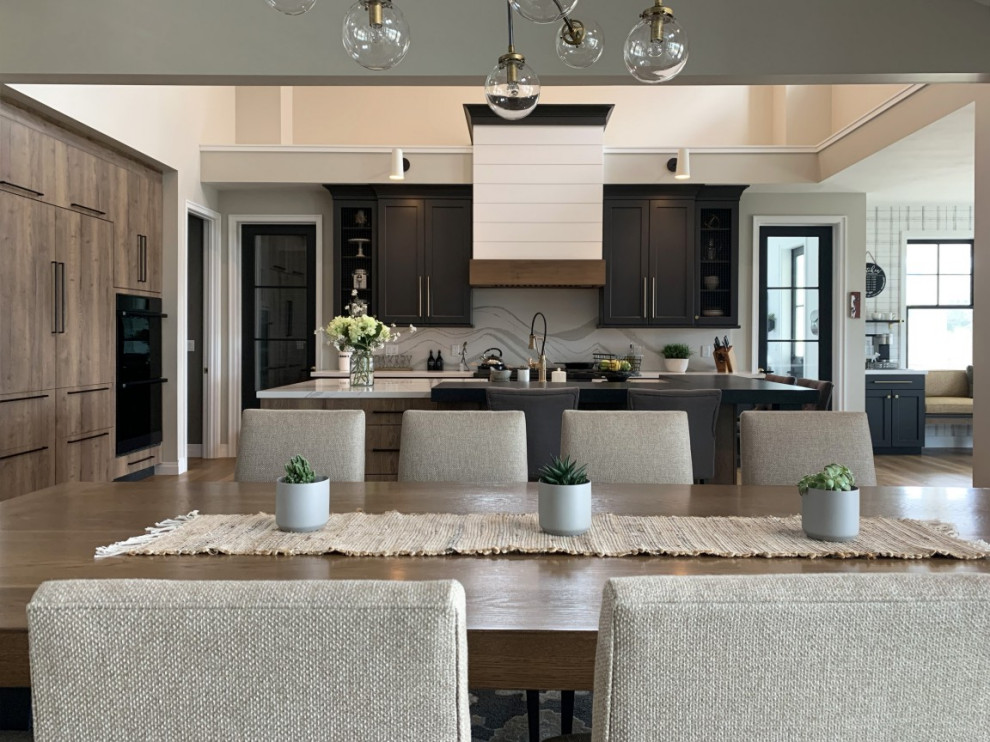 Transitional l-shaped light wood floor, brown floor and vaulted ceiling open concept kitchen photo in Santa Barbara with a farmhouse sink, flat-panel cabinets, distressed cabinets, quartz countertops, white backsplash, quartz backsplash, black appliances, an island and white countertops