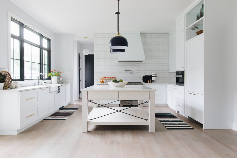 Design ideas for a country u-shaped kitchen in Chicago with a farmhouse sink, shaker cabinets, white cabinets, white splashback, subway tile splashback, panelled appliances, light hardwood floors, with island, beige floor and white benchtop.