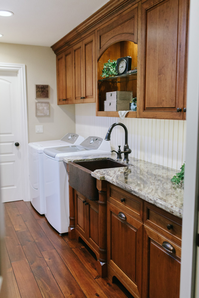 Mid-sized traditional single-wall dedicated laundry room in Sacramento with a farmhouse sink, raised-panel cabinets, medium wood cabinets, granite benchtops, white walls, medium hardwood floors and a side-by-side washer and dryer.
