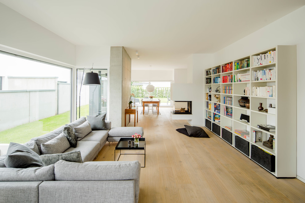 Large contemporary open concept living room in Dusseldorf with a library, white walls, light hardwood floors, a two-sided fireplace, a plaster fireplace surround and no tv.