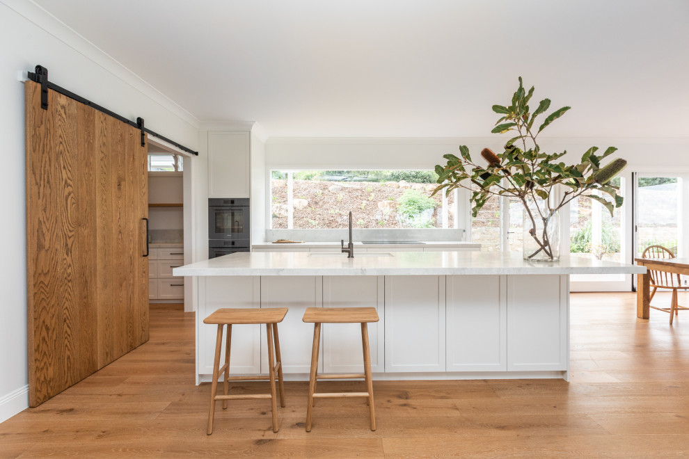 Design ideas for a large country open plan kitchen with an undermount sink, shaker cabinets, white cabinets, quartz benchtops, window splashback, light hardwood floors, with island and white benchtop.
