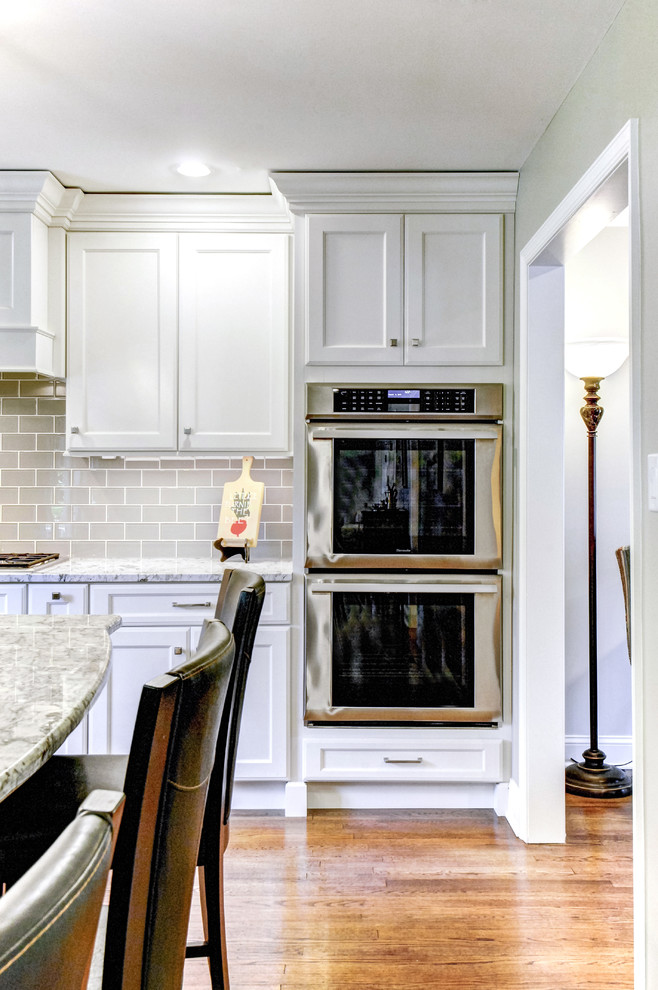 Photo of a large traditional l-shaped eat-in kitchen in Philadelphia with an undermount sink, recessed-panel cabinets, white cabinets, granite benchtops, white splashback, glass tile splashback, stainless steel appliances, medium hardwood floors and with island.