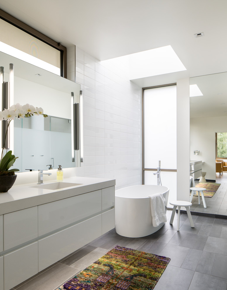 Photo of a contemporary bathroom in Denver with white cabinets, a freestanding tub, a curbless shower, a wall-mount toilet, white tile, ceramic tile, white walls, ceramic floors, an integrated sink, solid surface benchtops and flat-panel cabinets.