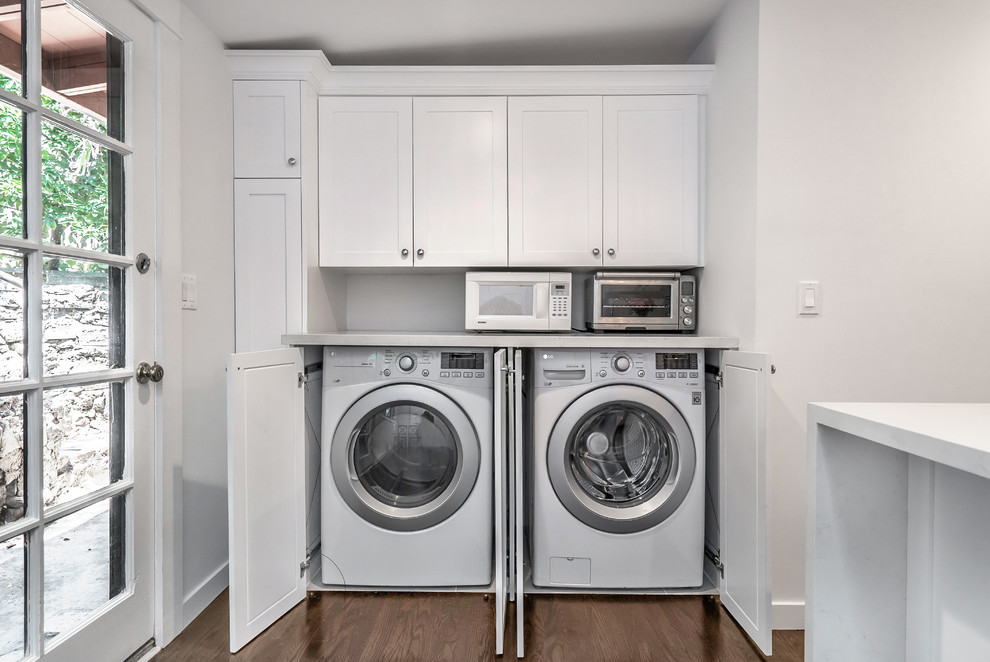 Photo of a mid-sized country u-shaped laundry room in Los Angeles with a farmhouse sink, shaker cabinets, white cabinets, quartz benchtops, white splashback, porcelain splashback, dark hardwood floors and brown floor.